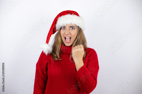 Young blonde woman wearing a red casual sweater and a christmas hat over white background angry and mad raising fist frustrated and furious while shouting with anger. Rage and aggressive concept.