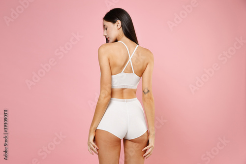 Back rear view of beautiful seductive sexy young brunette woman 20s in white underwear posing hold hands on hips legs looking down isolated on pastel pink colour background, studio portrait. © ViDi Studio
