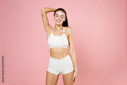 Smiling cute young brunette woman 20s in white underwear with fit sexy body posing put hands on head making ponytail keeping eyes closed isolated on pastel pink colour background, studio portrait.