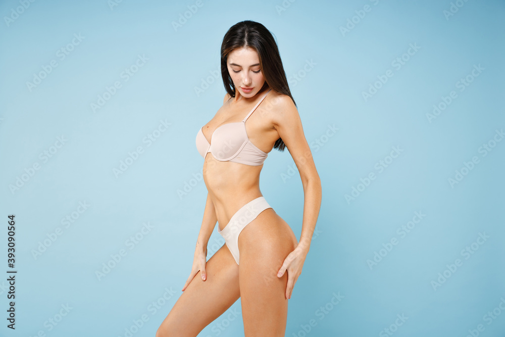 Side view of stunning attractive young brunette woman 20s in beige underwear showing sports body standing posing hold hand on hips legs isolated on pastel blue colour wall background studio portrait. Side view of stunning attractive young brunette woman 20s in beige underwear showing sports body standing posing hold hand on hips legs isolated on pastel blue colour wall background studio portrait.