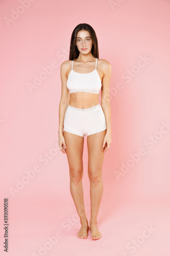 Full length of attractive beautiful fitness young brunette woman in white underwear showing fit sports body posing standing looking camera isolated on pastel pink colour background, studio portrait. © ViDi Studio