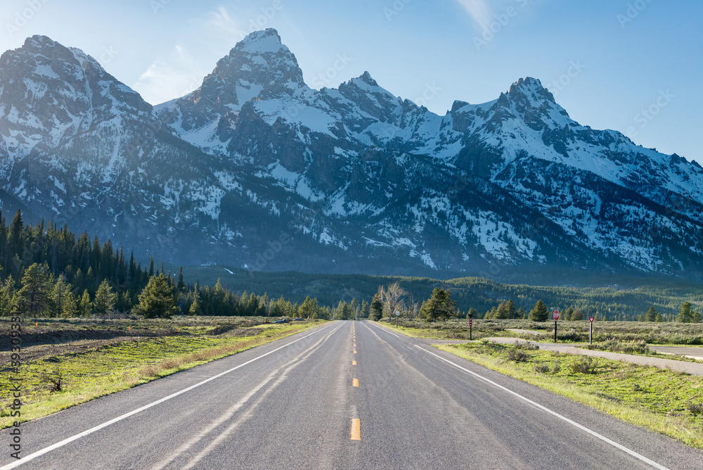 Straight road towards Grand Teton mountains at Grand Teton National Park, United States. Green tress and some light rays can be see coming from the left