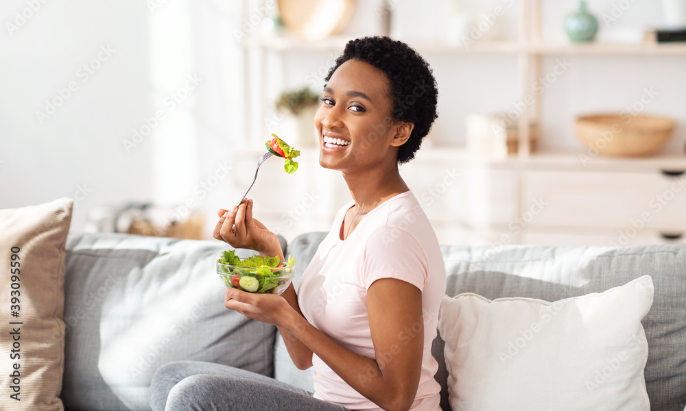 Healthy nutrition concept. Pretty black woman eating yummy vegetable salad on sofa at home, panorama