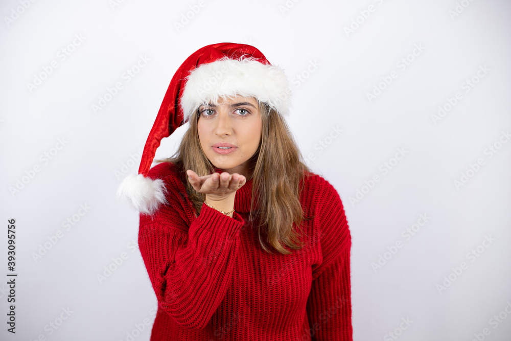 Young pretty blonde woman wearing a red casual sweater and a christmas hat over white background looking at the camera blowing a kiss with hand on air being lovely and sexy. Love expression.