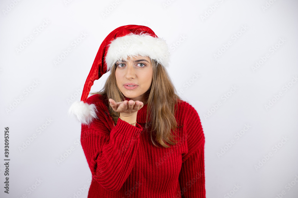 Young pretty blonde woman wearing a red casual sweater and a christmas hat over white background looking at the camera blowing a kiss with hand on air being lovely and sexy. Love expression. Young pretty blonde woman wearing a red casual sweater and a christmas hat over white background looking at the camera blowing a kiss with hand on air being lovely and sexy. Love expression.