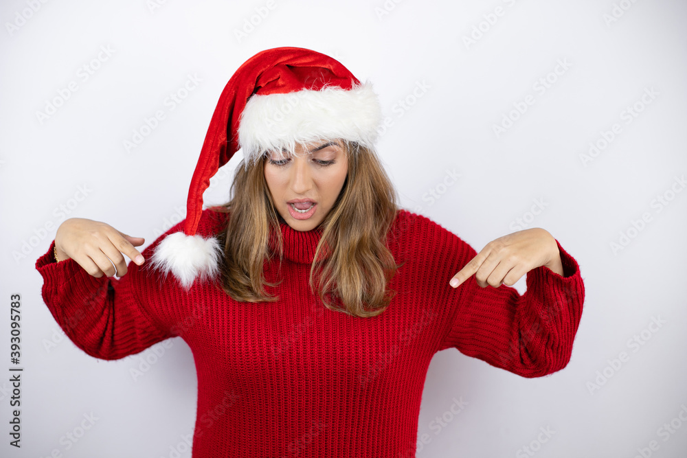 Young pretty blonde woman wearing a red casual sweater and a christmas hat over white background surprised, looking down and pointing down with fingers and raised arms Young pretty blonde woman wearing a red casual sweater and a christmas hat over white background surprised, looking down and pointing down with fingers and raised arms