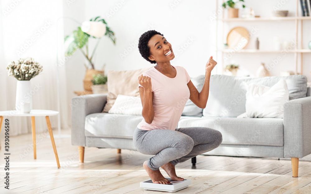 Excited black woman sitting on scales at home, happy with result of her slimming diet, copy space Excited black woman sitting on scales at home, happy with result of her slimming diet, copy space