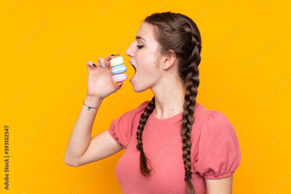 Young woman over isolated yellow background holding colorful French macarons and eating it Young woman over isolated yellow background holding colorful French macarons and eating it