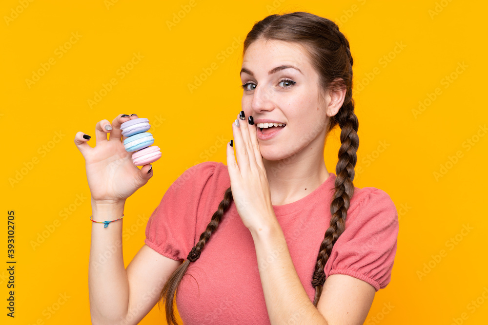 Young woman over isolated yellow background holding colorful French macarons and whispering something Young woman over isolated yellow background holding colorful French macarons and whispering something