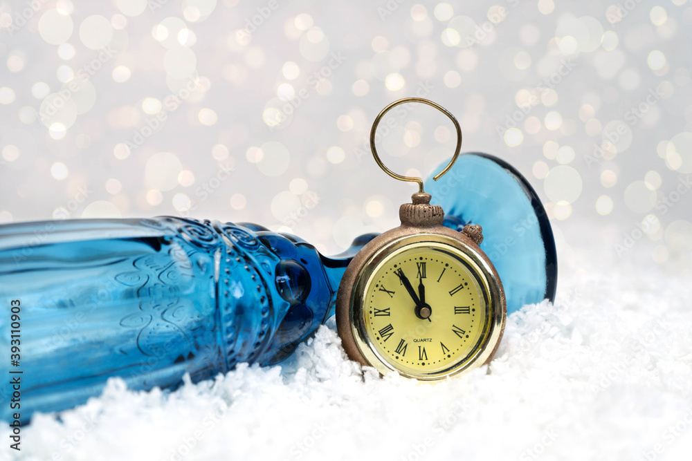 Christmas toy in the form of a clock and a blue champagne glass lie on the white snow. The gold clock shows five minutes to midnight. Bokeh in the background. Close-up