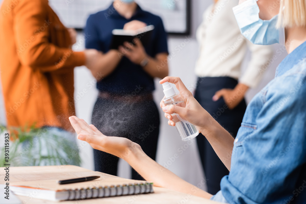 cropped view of businesswoman in medical mask spraying antiseptic on hands near colleagues on blurred background cropped view of businesswoman in medical mask spraying antiseptic on hands near colleagues on blurred background