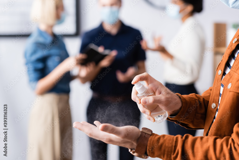 Obraz premium cropped view of businessman spraying sanitizer on hands while colleagues talking on blurred background