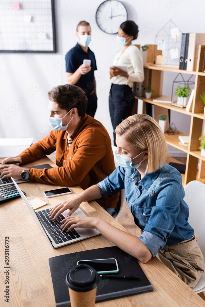 Fototapeta premium managers in medical masks working on laptops near multicultural colleagues talking on blurred background