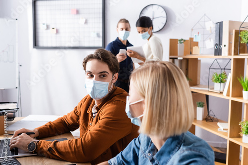young manager in medical mask looking at colleague while businesspeople talking on blurred background
