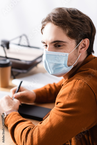 young manager in medical mask sitting at workplace with pen and notebook on blurred background