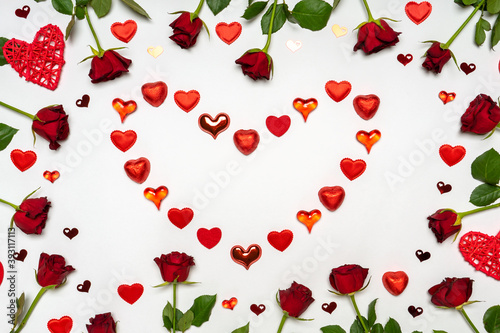 Heart symbol made of various red hearts and sweets with fresh red rose flowers on white background. Greeting card, mock up. Love, romance or Valentine's day concept. Flat lay, top view, copy space