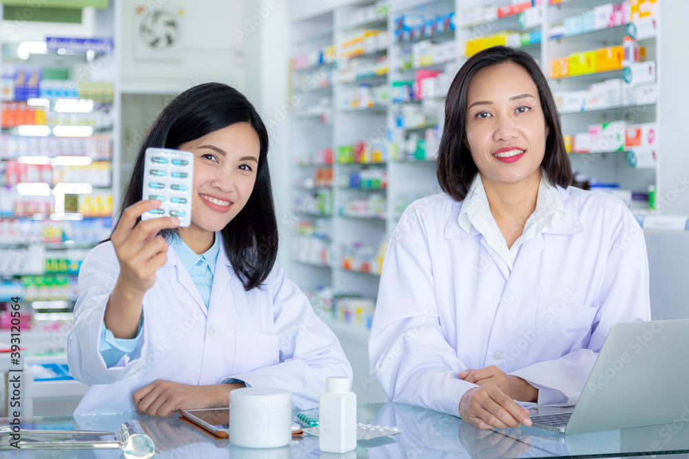Smiling asian pharmacist holding capsules medicine in pharmacy with her colleague. Healthcare and medical concept.