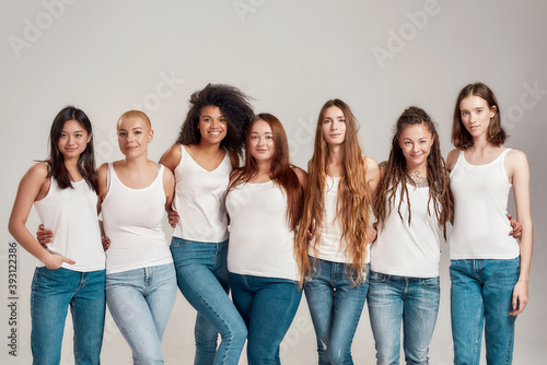 Group of beautiful diverse young women wearing white shirt and denim jeans looking at camera while posing together isolated over grey background