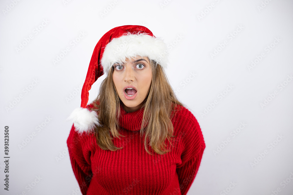 Young pretty blonde woman wearing a red casual sweater and a christmas hat over white background afraid and shocked with surprise and amazed expression, fear and excited face. Young pretty blonde woman wearing a red casual sweater and a christmas hat over white background afraid and shocked with surprise and amazed expression, fear and excited face.