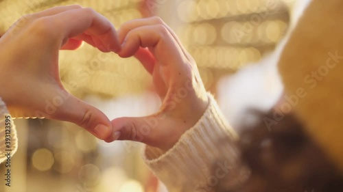 Young woman makes heart with palms against blurred colorful glowing marry-go-round in contemporary amusement park at sunset extreme close view photo