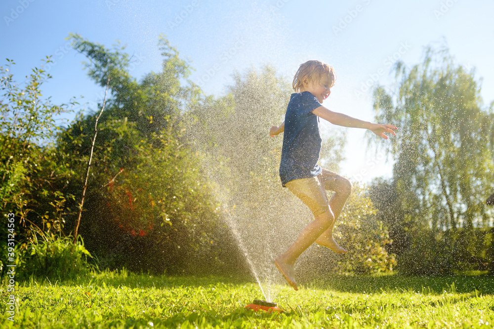 Funny little boy playing with garden sprinkler in sunny backyard. Preschooler child laughing, jumping and having fun with spray of water. - obrazy, fototapety, plakaty 