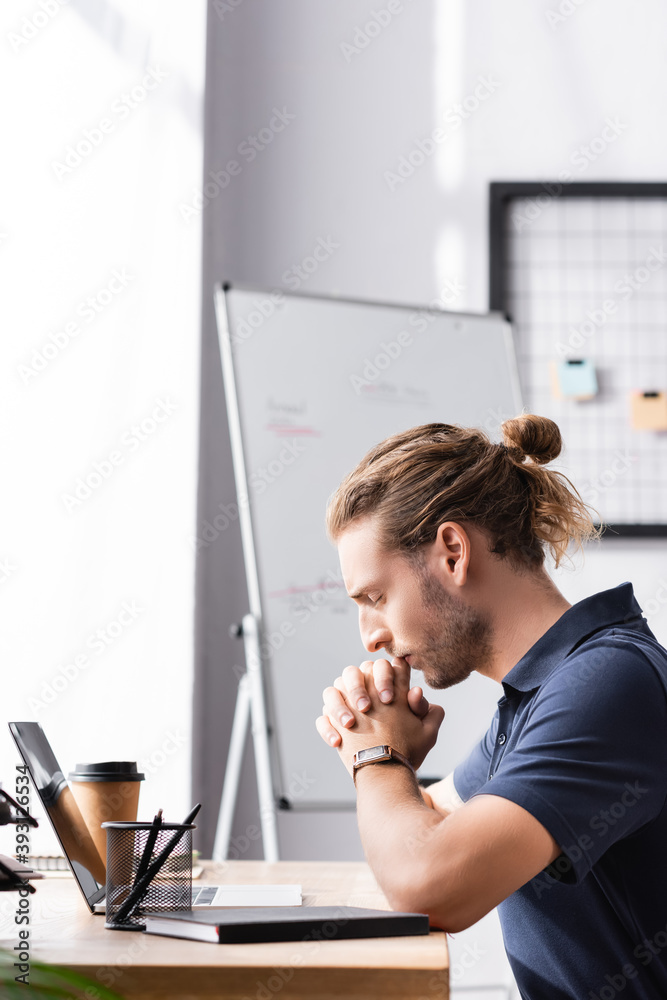 Thoughtful office worker with clenched hands sitting at workplace table near laptop on blurred background