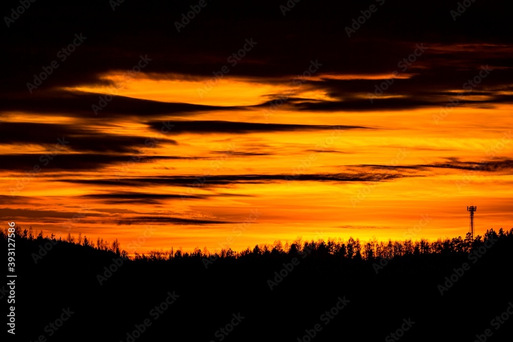 Bright orange and yellow sunset sky with clouds and black horizon with trees and a silhouette of the communication tower.