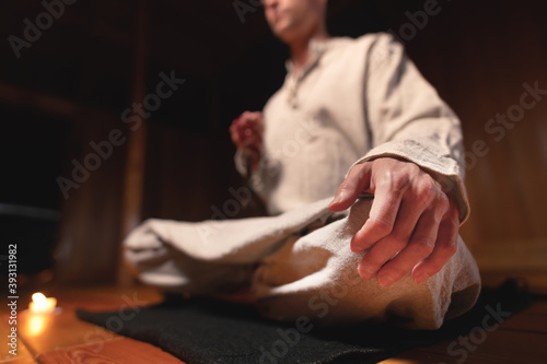 A young man in gray clothes sits in a dark room in a lotus position with a rosary in his hands, surrounded by candles. Practice and religion