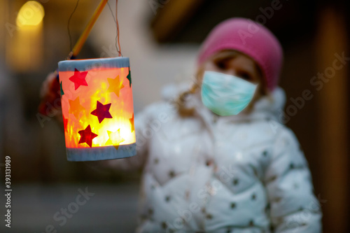 Toddler girl holding selfmade lantern for St. Martin procession. Child wear medical mask. children and family parade in kindergarten. German tradition Martinsumzug Pandemic corona virus quarantine