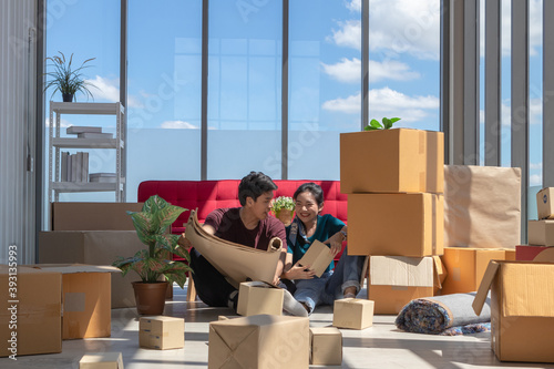 Young Asian couple relax together after moving house. Lean against red sofa with many boxes in living room. Talking or discuss with house plan and design.