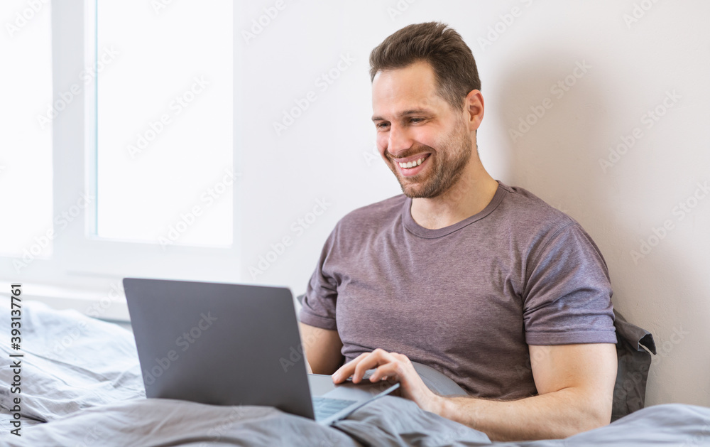 Cheerful Guy Using Laptop Working Online Remotely Sitting In Bedroom Cheerful Guy Using Laptop Working Online Remotely Sitting In Bedroom