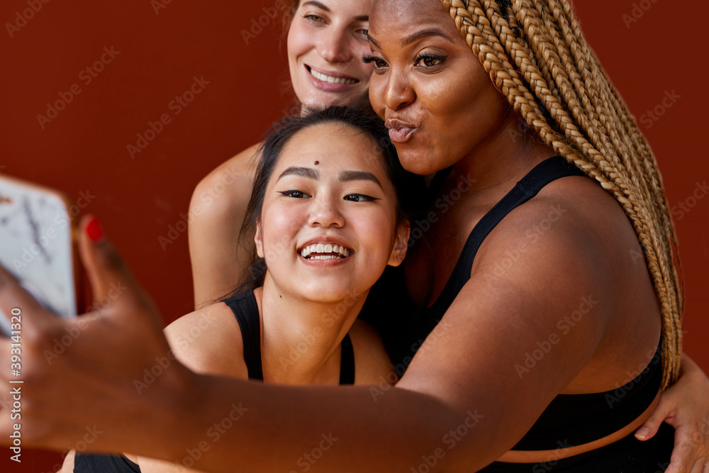 three young multi ethnic female models of different race, hair colour and body size taking selfie at camera isolated on red background, have fun, they are in black sportswear three young multi ethnic female models of different race, hair colour and body size taking selfie at camera isolated on red background, have fun, they are in black sportswear