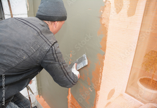 A handyman is skim coating  plastering  finishing  rendering the exterior wall  and applying cement over fiberglass reinforcing mesh around the window area on the outside wall of the building.