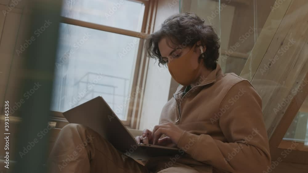Low angle view of young curly-haired Caucasian man wearing casual clothes and protective mask sitting on wooden stairs, working on laptop and then looking at camera Low angle view of young curly-haired Caucasian man wearing casual clothes and protective mask sitting on wooden stairs, working on laptop and then looking at camera