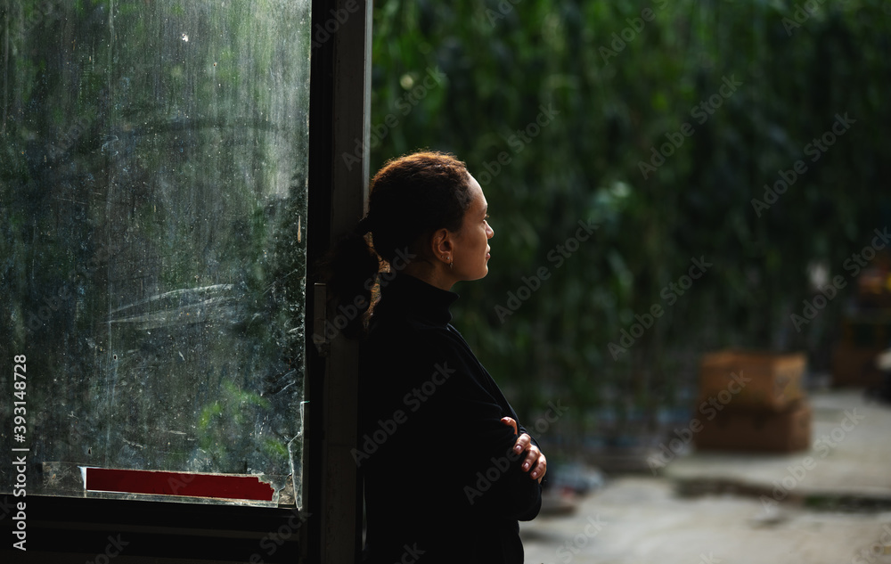 Business in modern greenhouses all year round. Side view of a young female entrepreneur standing at the entrance to a hangar. Green plants are visible in the background. Business in modern greenhouses all year round. Side view of a young female entrepreneur standing at the entrance to a hangar. Green plants are visible in the background.