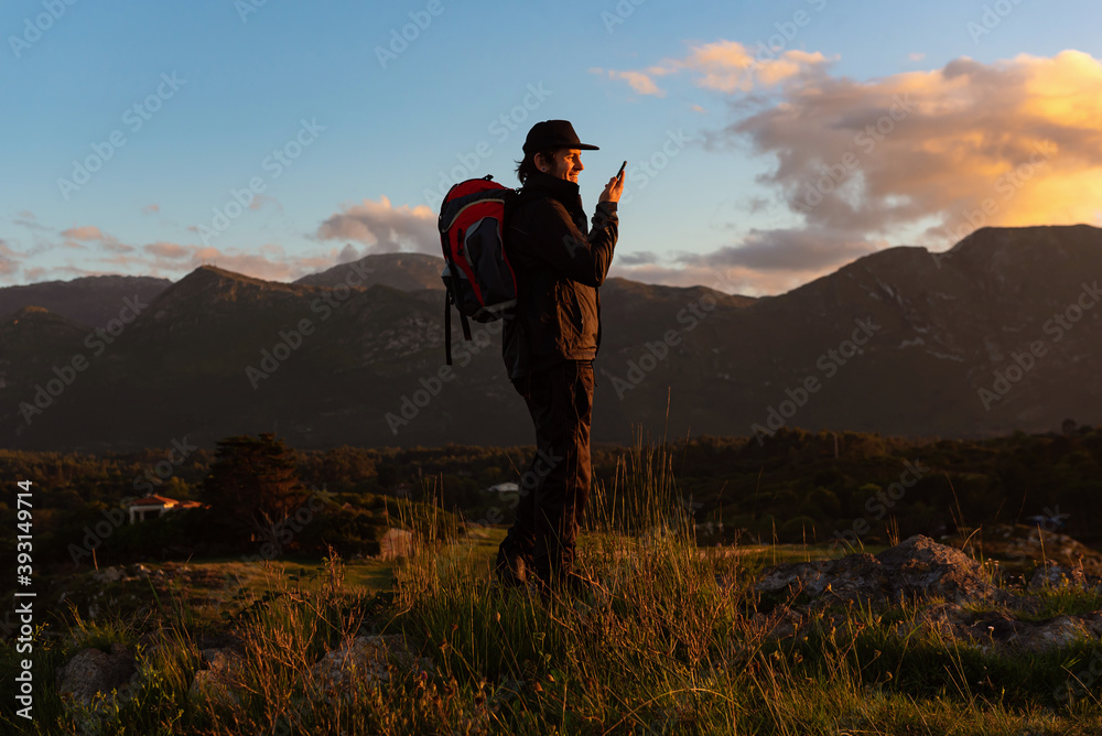 Side view of hiker standing on rock in highlands and speaking on cellphone while admiring sunset Side view of hiker standing on rock in highlands and speaking on cellphone while admiring sunset