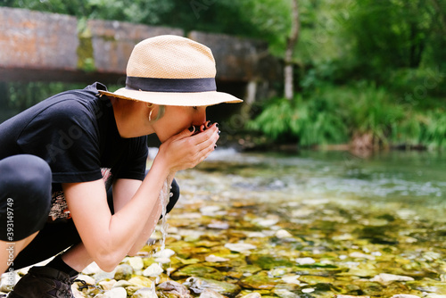 Side view of female tourist drinking clear water from calm river in forest during summer adventure