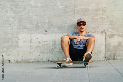 Confident male skater in stylish outfit sitting near urban building with skateboard and looking at camera photo
