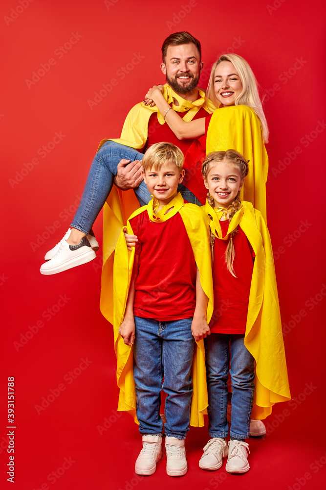 family pretending to be superhero in the park, beautiful happy parents and children in superhero costume posing isolated over red background. entertainment concept