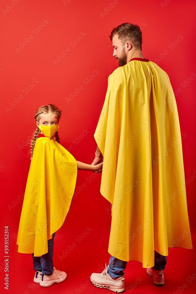 father and daughter ready to fight against coronavirus, they stand in superhero costume, girl in medical mask, look at camera. isolated over red background