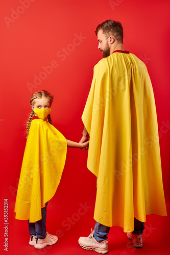 father and daughter ready to fight against coronavirus, they stand in superhero costume, girl in medical mask, look at camera. isolated over red background