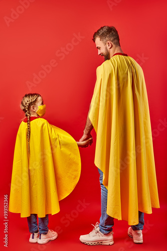 friendly man and kid girl in yellow cloaks, pretend to be superheroes. male and child in mask stand holding hands together, rear view