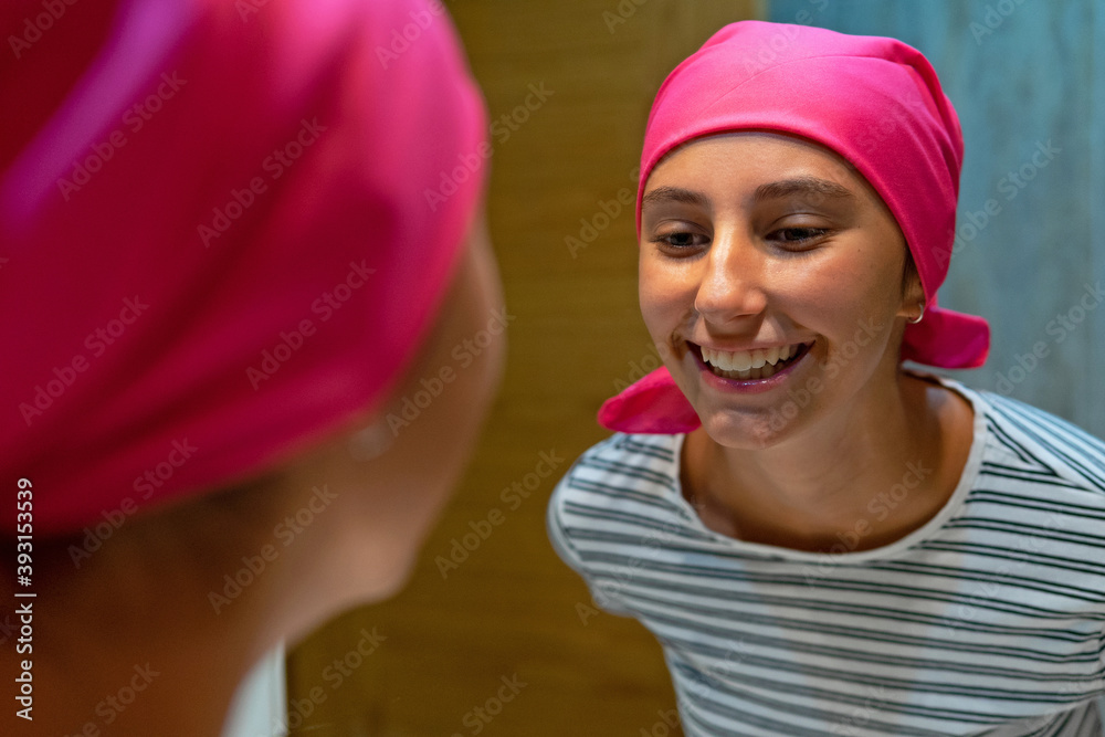Optimistic young female with cancer and in headscarf standing at home and charmingly looking in mirror Optimistic young female with cancer and in headscarf standing at home and charmingly looking in mirror