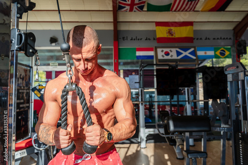 Powerful male athlete with sweaty naked torso pulling ropes of cable machine and pumping biceps while training in modern gym