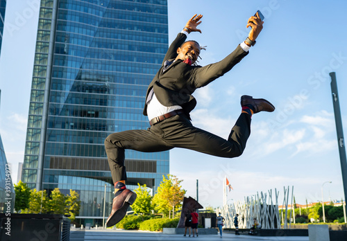 Side view of carefree black male entrepreneur in classy suit and with smartphone jumping on street in downtown and looking away photo