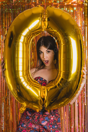 Happy young female holding bright golden balloon in shape of number 0 while standing against wall with glowing multicolored tinsel during New Year celebration