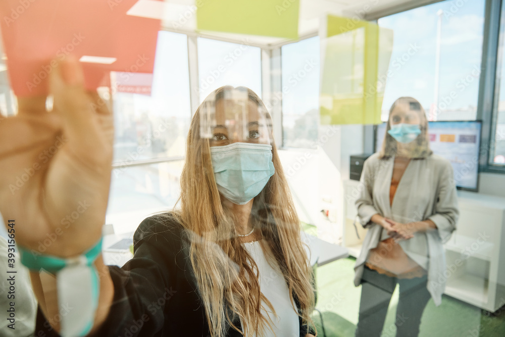 © Philippe Degroote/ADDICTIVE STOCK - Through glass view of young creative female coworkers in medical masks working with sticky notes while discussing marketing strategy during meeting in contemporary workspace