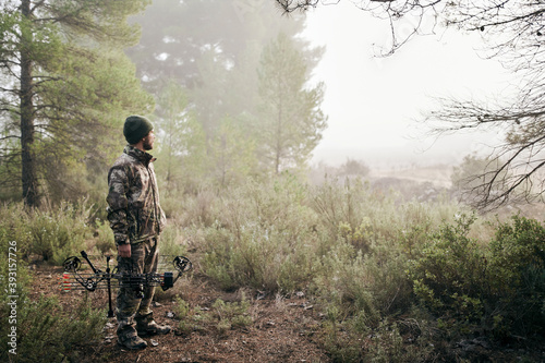 Side view of man in camouflage standing with compound bow in forest and looking away during hunting