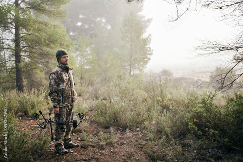Side view of man in camouflage standing with compound bow in forest and looking away during hunting photo