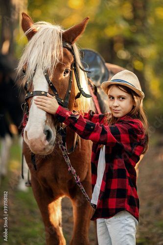 a teenage girl in a plaid shirt and hat holds the reins of a brown horse on an autumn farm